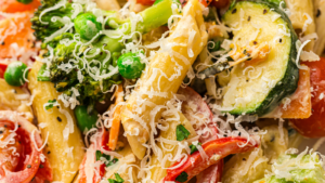 Close-up of penne pasta mixed with broccoli, peas, zucchini, red bell pepper, and topped with grated cheese.
