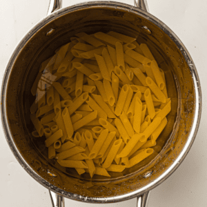A stainless steel pot filled with uncooked penne pasta soaking in water, viewed from above against a white background.