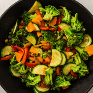 A cast iron pan filled with stir-fried vegetables, including broccoli, zucchini, carrots, peas, and red bell peppers.