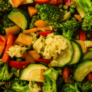 Close-up of assorted stir-fried vegetables including broccoli, zucchini, carrots, red bell pepper, peas, and a topping of minced garlic.