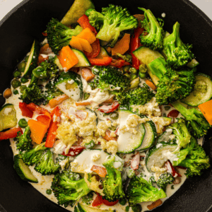 A close-up of a skillet filled with broccoli, zucchini, carrots, peas, and red peppers cooking in a creamy sauce.