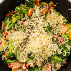 A skillet filled with mixed vegetables such as broccoli, zucchini, and bell peppers, topped with shredded cheese and ground black pepper.