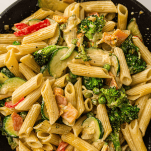 Penne pasta mixed with broccoli, zucchini, red bell pepper, peas, and a creamy sauce, served in a black bowl.