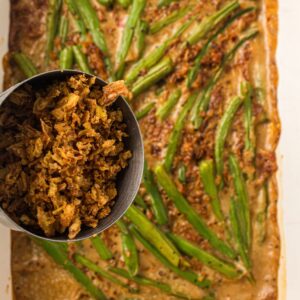 A metal cup filled with crispy fried onions is held over a rectangular flatbread topped with sliced green vegetables and sauce.