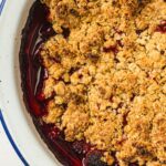 A close-up of a baked strawberry crumble dessert with a golden oat topping and visible red fruit filling in a white dish.