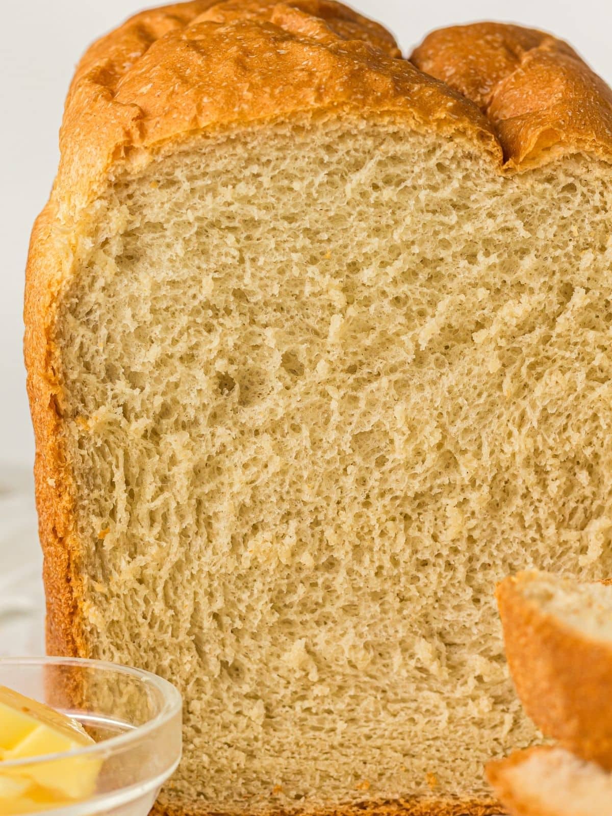 Close-up of a loaf of bread with a golden crust, showing its soft, airy texture inside. A small bowl with butter and a sliced piece of bread are visible in the foreground.