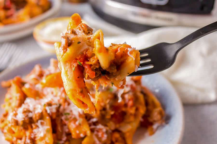 A black fork holds a bite of cheesy pasta with meat sauce above a plate filled with more pasta.