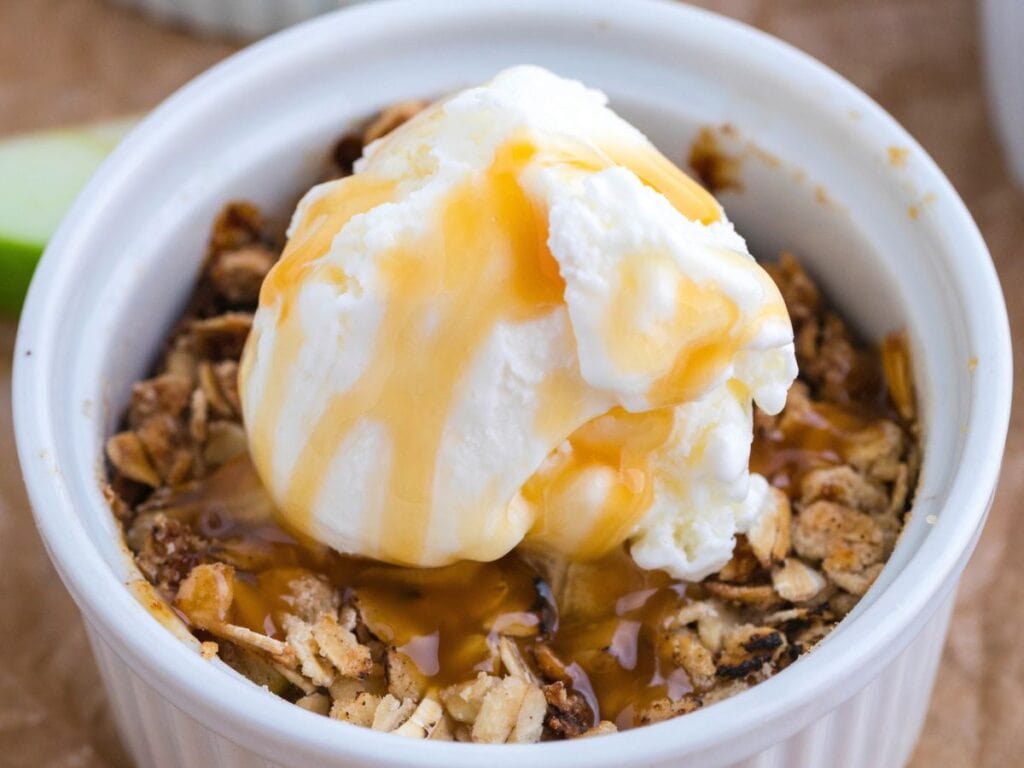 A white ramekin filled with apple oat crumble, topped with a scoop of vanilla ice cream and drizzled with caramel sauce.