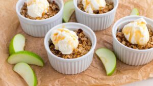 Four ramekins filled with apple crisp topped with ice cream and caramel sauce, placed on brown parchment paper with green apple slices.