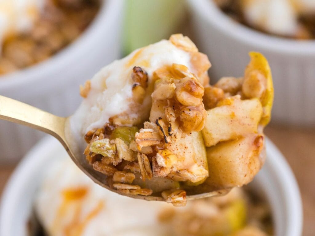 A close-up of a spoonful of apple crisp with oats and a dollop of whipped cream, held above a white ramekin.