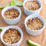 Four white ramekins filled with baked oat and apple crumble are arranged on brown parchment paper, with green apple slices placed around them.