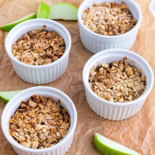 Four white ramekins filled with baked oat and apple crumble are arranged on brown parchment paper, with green apple slices placed around them.