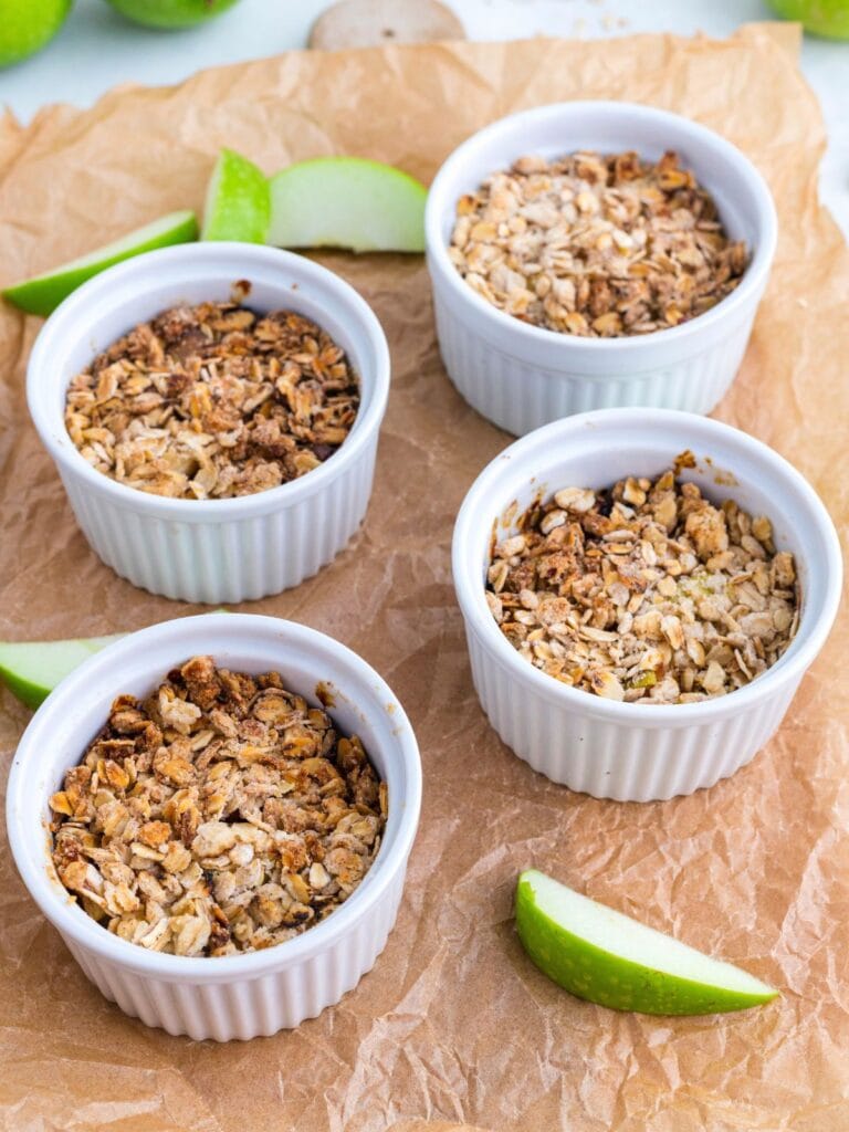 Four white ramekins filled with baked oat and apple crumble are arranged on brown parchment paper, with green apple slices placed around them.