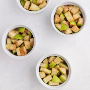 Four white ramekins filled with chopped green apples sprinkled with cinnamon and sugar, arranged on a white surface.