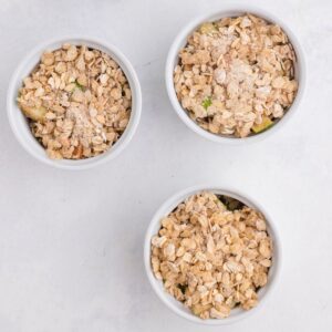 Three white ramekins filled with an oat crumble topping, arranged in a triangle on a light-colored surface.