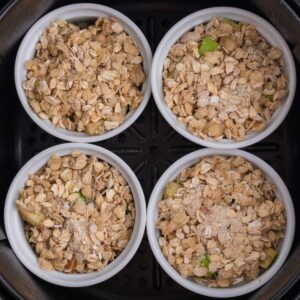 Four white ramekins filled with oat crumble topping and diced fruit are arranged in a black air fryer basket, ready to be baked.
