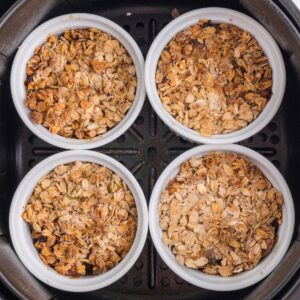 Four white ramekins filled with baked oat crumble sit inside a black air fryer basket, viewed from above.