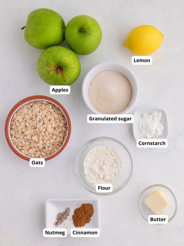 Flat lay of baking ingredients: three green apples, a lemon, granulated sugar, cornstarch, oats, flour, butter, ground cinnamon, and ground nutmeg on a white surface.