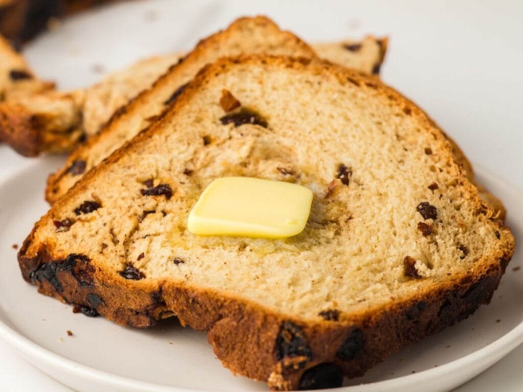 Two slices of raisin bread on a white plate, with a pat of butter melting on the top slice.