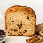 A loaf of raisin bread is sliced open, showing its texture and raisins inside. Cinnamon sticks and a small bowl of raisins are placed next to the bread on a white surface.