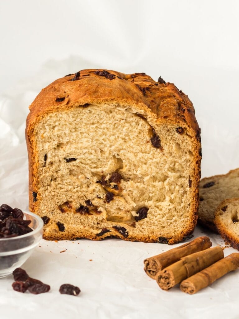 A loaf of raisin bread is sliced open, showing its texture and raisins inside. Cinnamon sticks and a small bowl of raisins are placed next to the bread on a white surface.