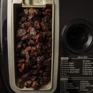 A close-up of a bread machine’s raisin dispenser filled with raisins, next to a control panel with a menu of baking options.
