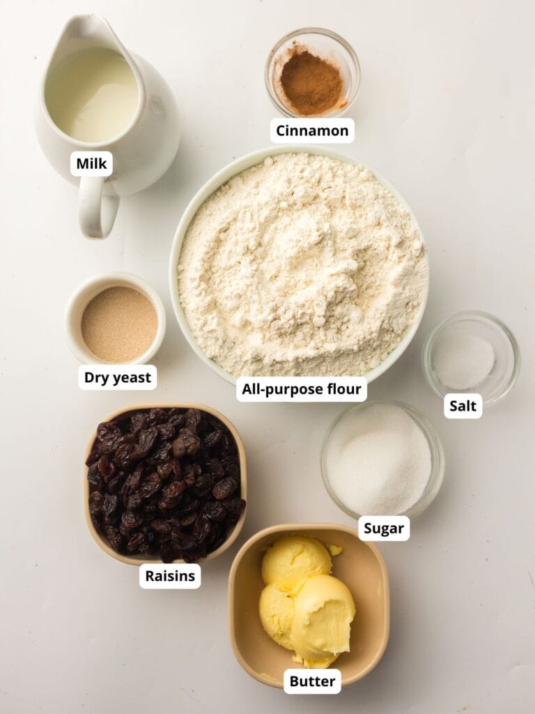 Various baking ingredients, including milk, cinnamon, all-purpose flour, dry yeast, salt, sugar, raisins, and butter, arranged in separate bowls on a white surface.