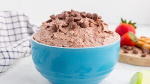 A blue bowl filled with chocolate chip-topped whipped dessert sits on a white surface with fruit and a cloth in the background.