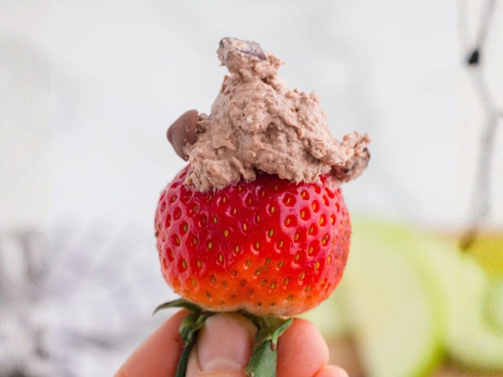 A hand holds a fresh strawberry topped with a dollop of chocolate whipped cream.