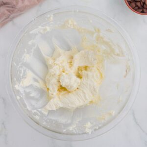 A glass bowl containing a mixture of creamed butter and sugar sits on a white surface, alongside a bowl of chocolate chips in the background.