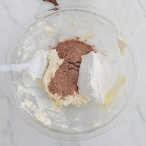 A glass bowl with whipped cream, chocolate powder, and butter mixture being mixed with a white spatula on a marble surface.
