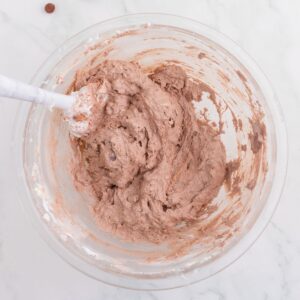 A clear bowl containing chocolate cookie dough being mixed with a white spatula on a marble countertop.