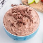 A blue bowl filled with chocolate-flavored whipped dip, topped with chocolate chips. Sliced green apples and a wooden board are partially visible in the background.