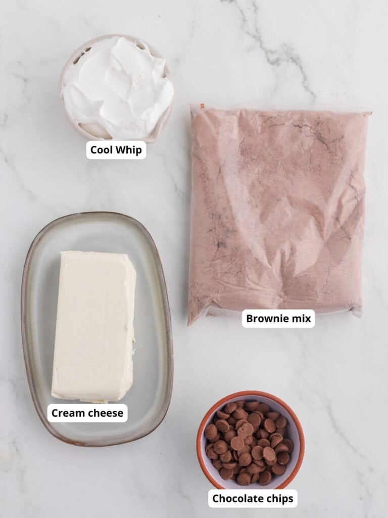 Four ingredients on a marble surface: a tub of Cool Whip, a bag of brownie mix, a block of cream cheese, and a small bowl of chocolate chips.