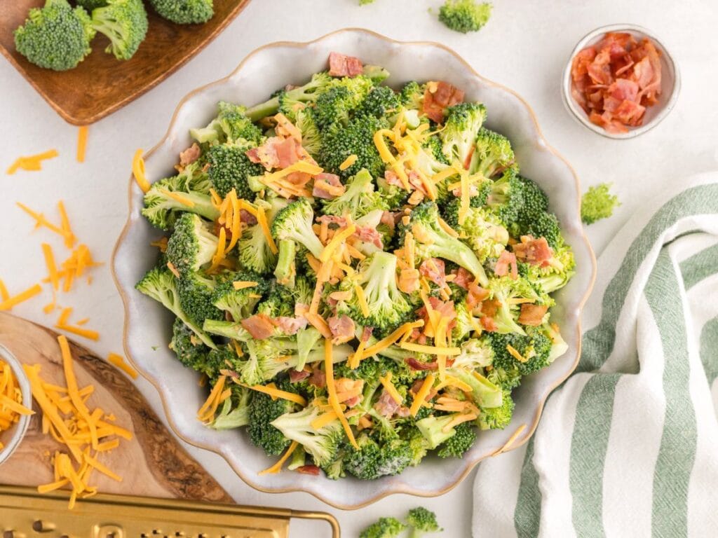Bowl of broccoli salad topped with shredded cheddar cheese and bacon bits, with ingredients and a grater nearby on a white surface.