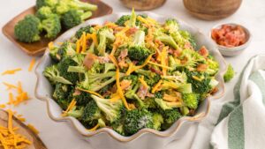 A bowl of broccoli salad topped with shredded cheddar cheese and bacon bits, surrounded by small dishes of broccoli florets and chopped ingredients.