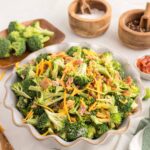 A bowl of broccoli salad with shredded cheese, bacon, and dressing, surrounded by bowls of salt, pepper, chopped tomatoes, and fresh broccoli on a white countertop.