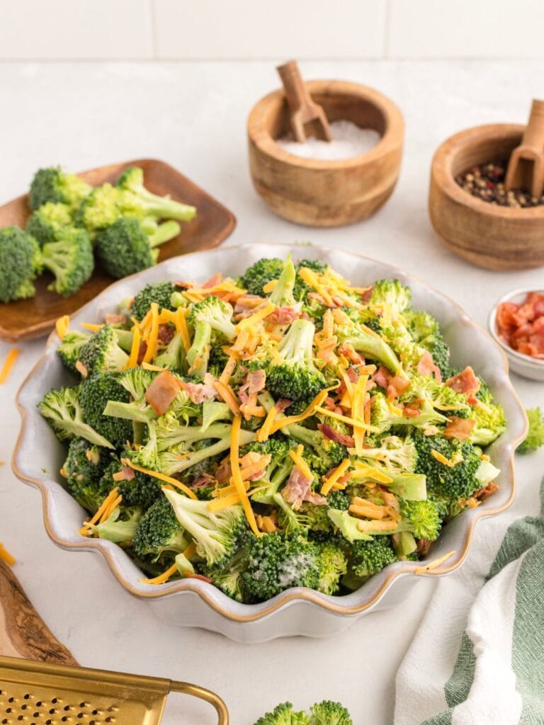 A bowl of broccoli salad with shredded cheese, bacon, and dressing, surrounded by bowls of salt, pepper, chopped tomatoes, and fresh broccoli on a white countertop.