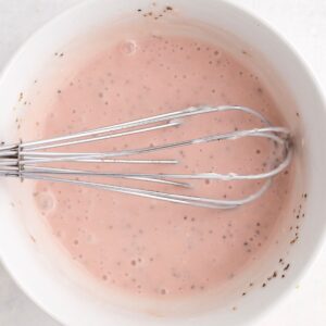 A metal whisk rests in a white bowl filled with pink batter or sauce, speckled with small dark bits.