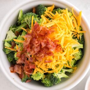 A white bowl filled with chopped broccoli, shredded cheddar cheese, and crumbled cooked bacon.