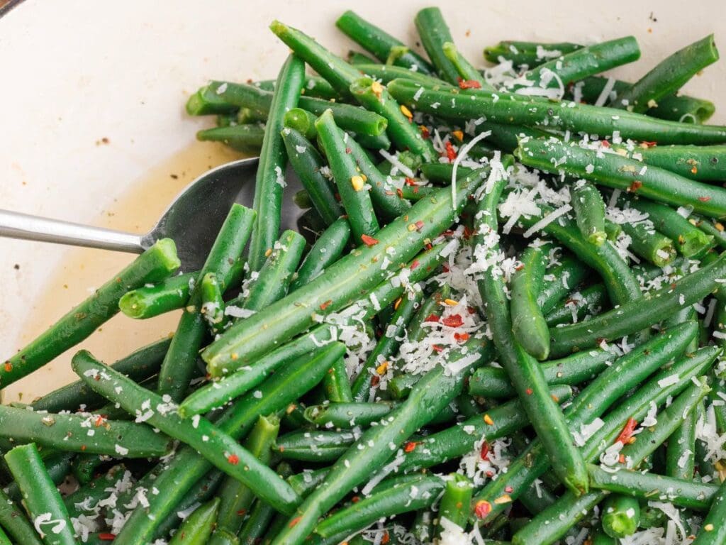 Cooked green beans topped with grated cheese and red pepper flakes, served in a white dish with a metal serving spoon.