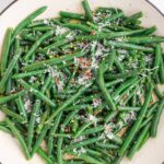 A white dish filled with cooked green beans topped with grated cheese and red pepper flakes, viewed from above.