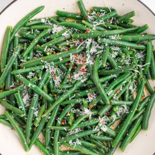 A white dish filled with cooked green beans topped with grated cheese and red pepper flakes, viewed from above.
