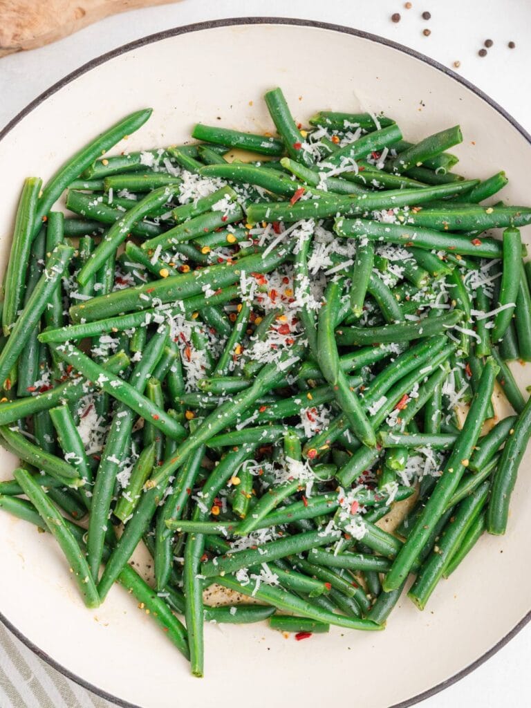A white dish filled with cooked green beans topped with grated cheese and red pepper flakes, viewed from above.