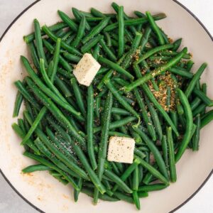 Fresh green beans in a pan with two cubes of butter and sprinkled seasonings, ready to be cooked.