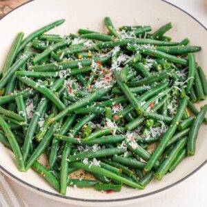 A shallow white bowl filled with cooked green beans, topped with grated cheese and red pepper flakes.