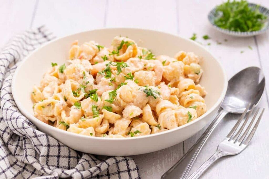 A bowl of creamy pasta with chopped herbs on top, placed on a striped napkin next to a fork and spoon on a white wooden table.