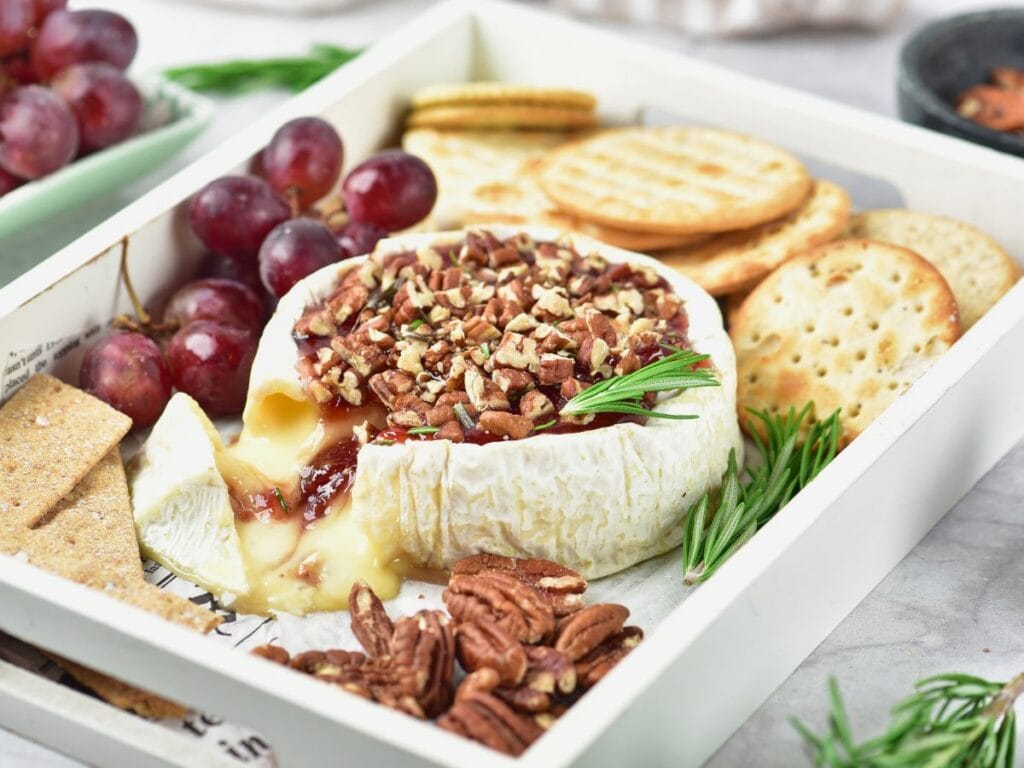 Baked brie topped with jam and pecans, served on a tray with grapes, crackers, and sprigs of rosemary.