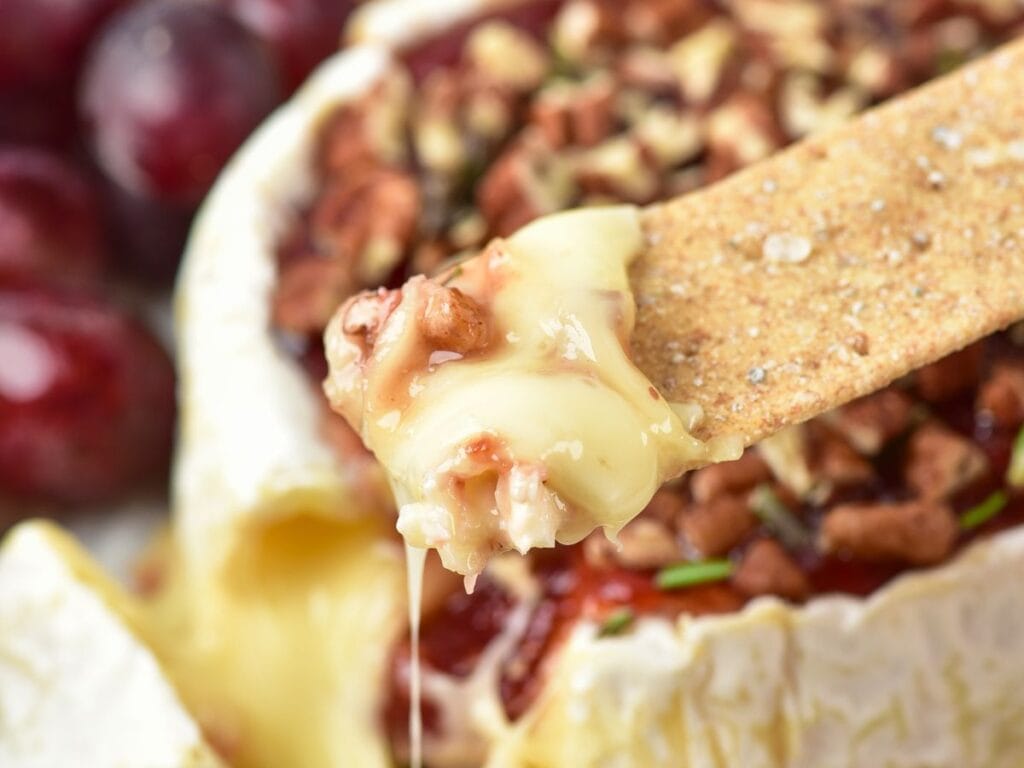 A cracker lifting a portion of melted brie topped with pecans and red preserves, with grapes in the background.