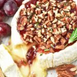 Baked Brie topped with red jam, chopped pecans, and rosemary sprigs, with grapes and crackers nearby.